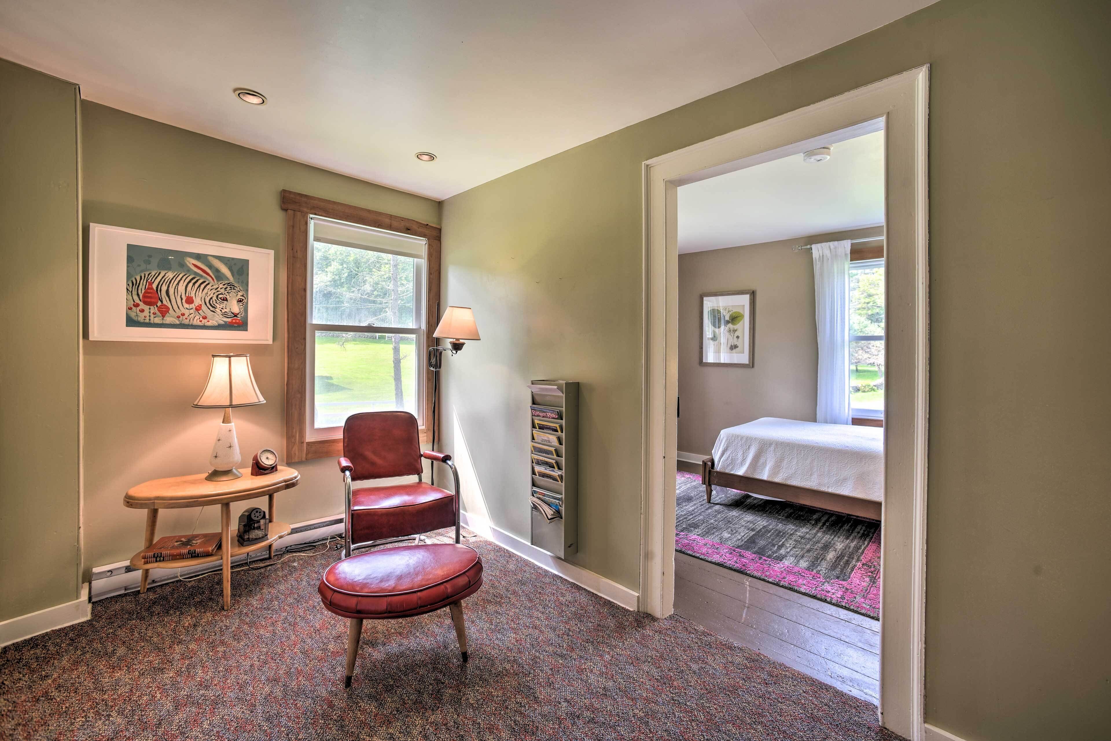A room with a red leather chair, a small table with a lamp, a bookshelf, and a bed with a white bedspread.