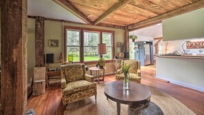 Ein rustikales Interieur mit Holzbalken, einer Essecke mit rundem Tisch, zwei gemusterten Sesseln und Blick ins Freie durch große Fenster.