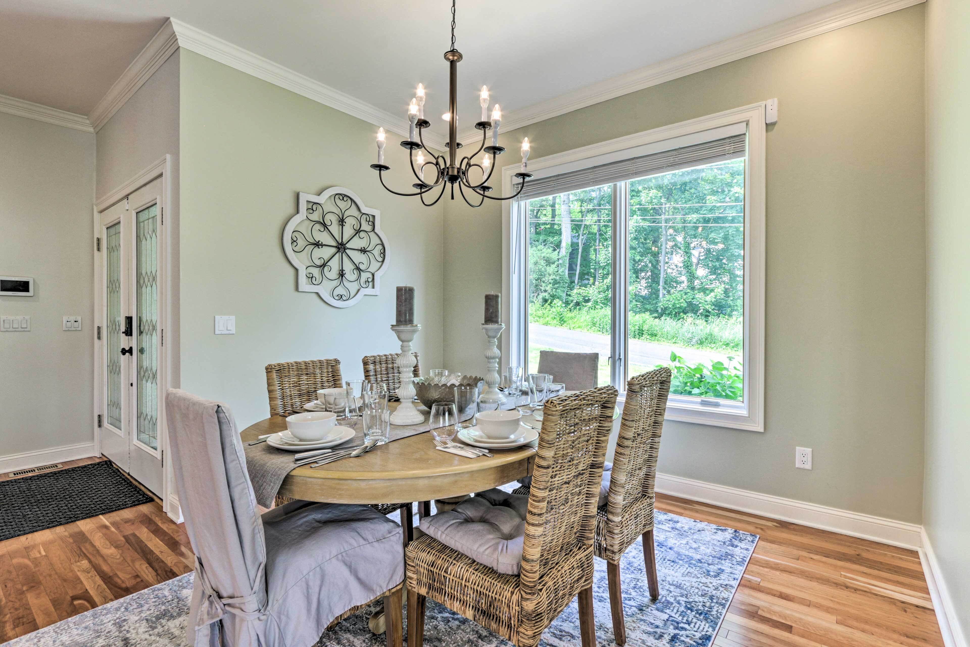 A dining room with a round table set for six, surrounded by wicker chairs, a chandelier, and a large window with a view of greenery.