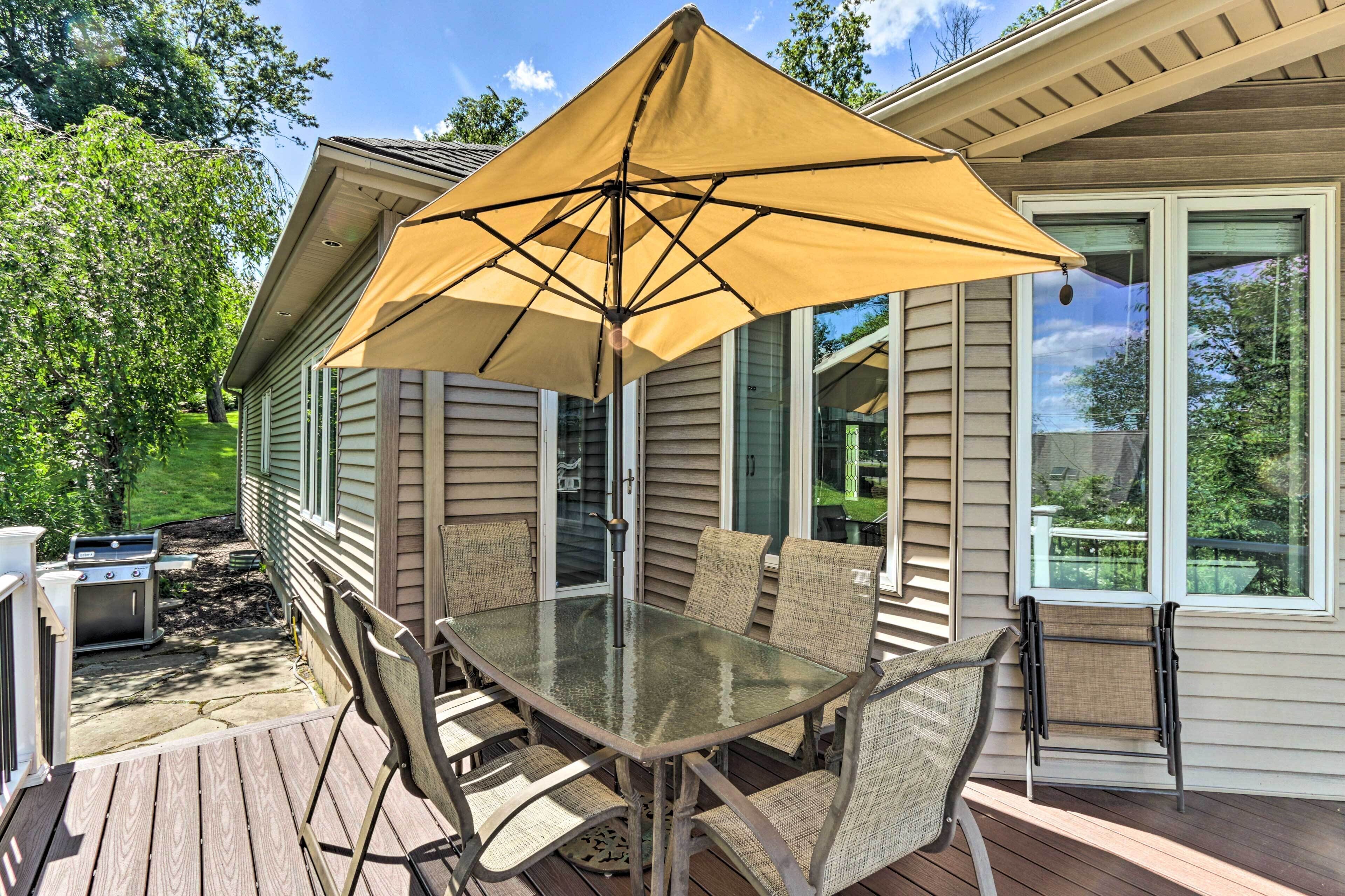 Een terras buiten met een glazen tafel en stoelen, een gele parasol en een barbecue.