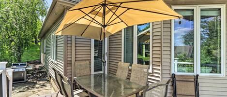 Een terras buiten met een glazen tafel en stoelen, een gele parasol en een barbecue.