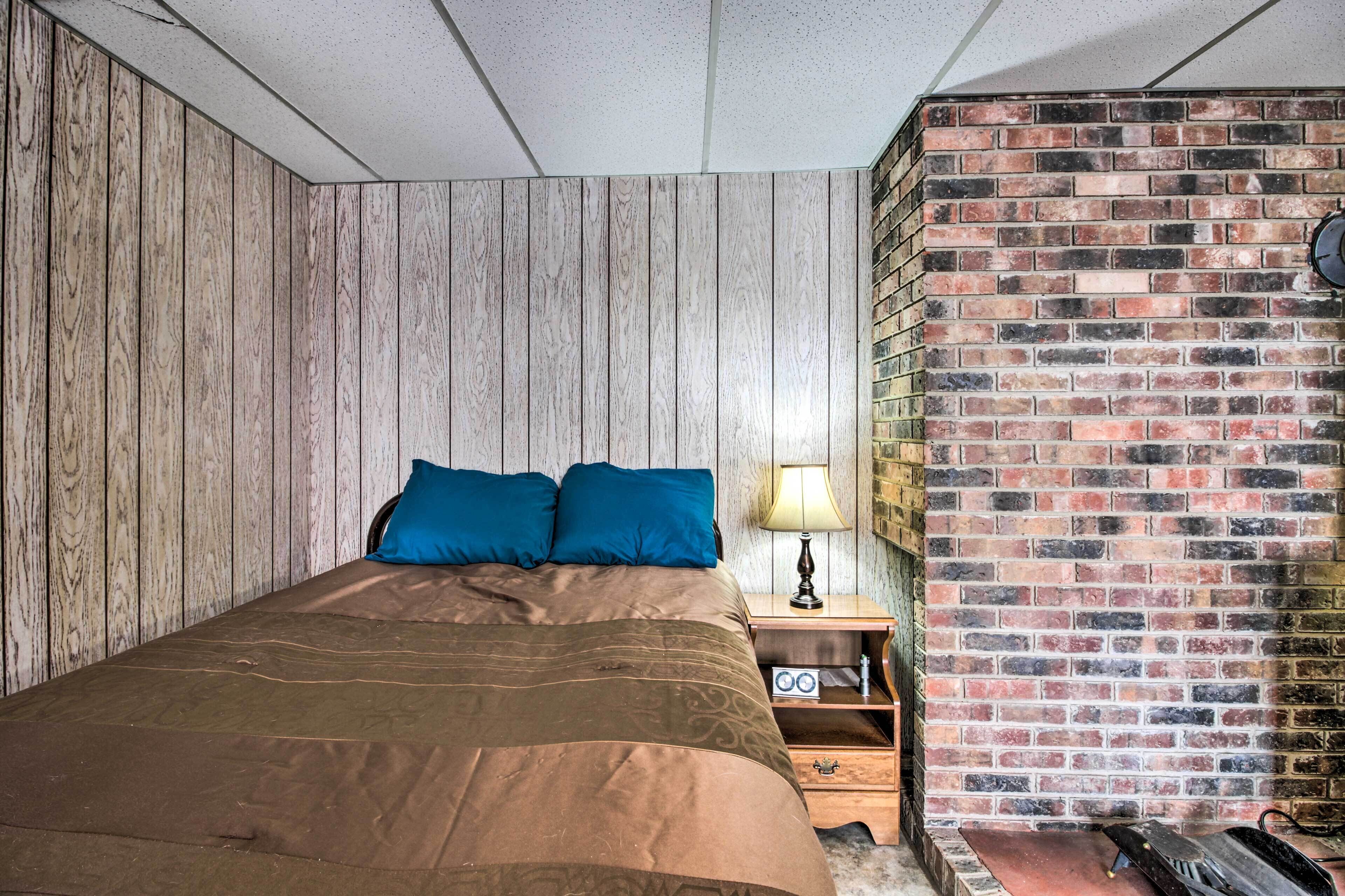 A bedroom with a wooden wall, a brick wall, a bed with blue pillows, a nightstand with a lamp, and a radio.