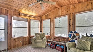 Interno accogliente di una baita con pareti in legno, ampie finestre, una ventola a soffitto e un'area salotto confortevole con divano e poltrona.
