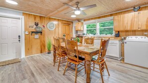 A wooden cabin interior with a dining table, chairs, a ceiling fan, a clock, and a kitchen area with cabinets and a stove. - 2 Mi to Ski Sapphire: Pet-friendly Cashiers Cabin (Cashiers)