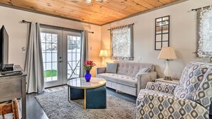 A cozy living room with a wooden ceiling, a grey sofa, a patterned armchair, a round coffee table with a vase of flowers, and a lamp on a side table. - Bemus Point Vintage Suite w/ Soaking Tub & Kitchen (Bemus Point)
