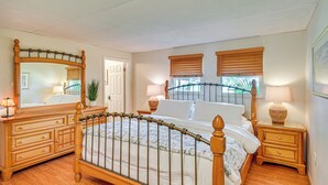 Une chambre à coucher avec un lit en bois, une commode, une table de chevet, un miroir et deux fenêtres équipées de stores.