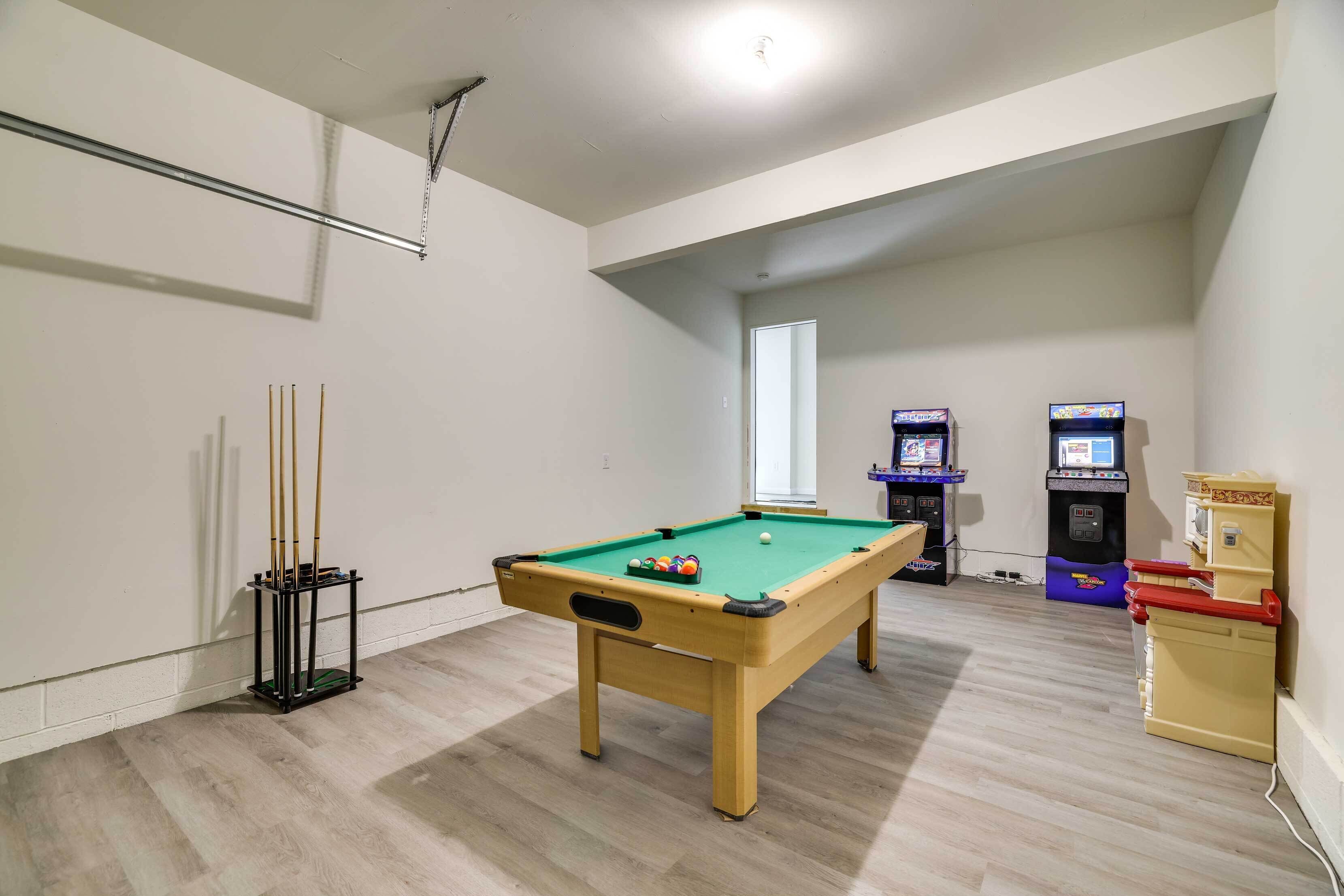 A pool table with a green felt surface, a pool cue rack, and two arcade machines in a room with wooden flooring and white walls.