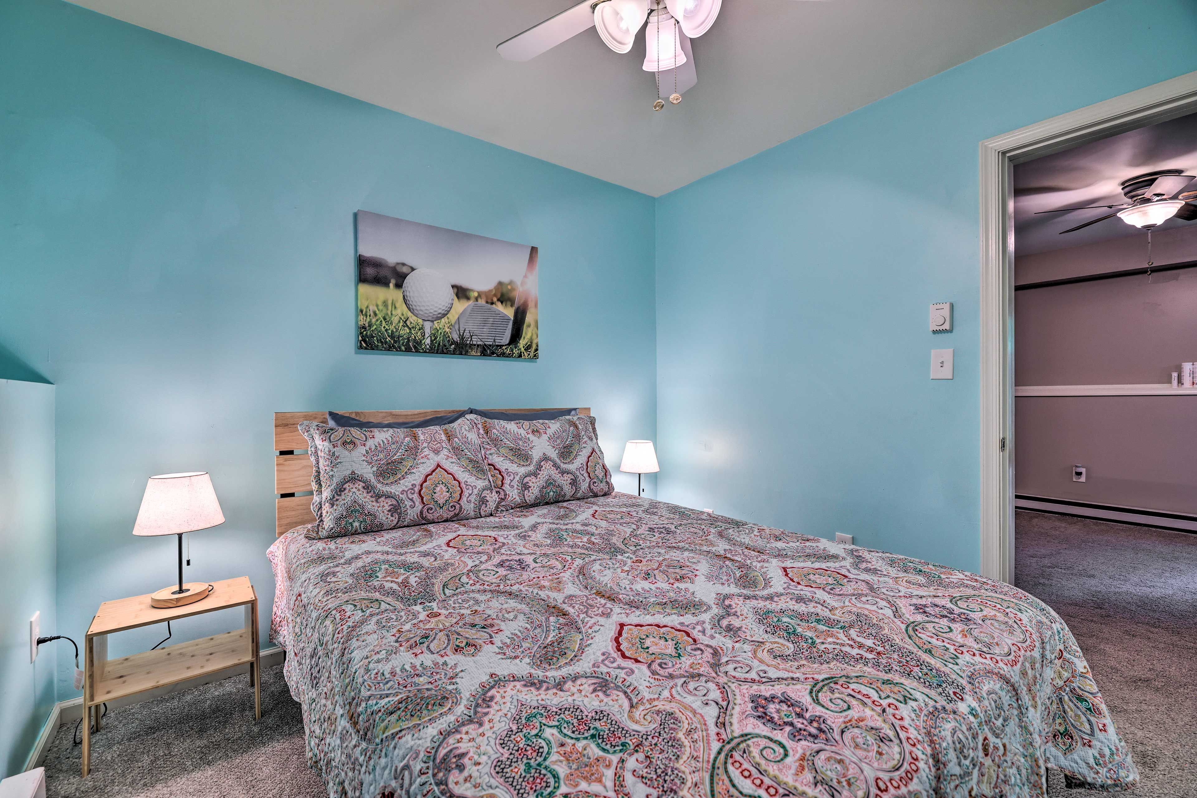 Une chambre à coucher comprenant un lit, une table de chevet, une lampe, un ventilateur de plafond et un tableau accroché au mur.