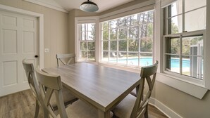 Un coin repas avec une table et des chaises en bois, de grandes fenêtres donnant sur une piscine et des arbres, et une porte blanche.