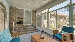 Une véranda avec un mur en briques, de grandes fenêtres, un ventilateur de plafond, une table en bois et deux chaises avec des coussins.