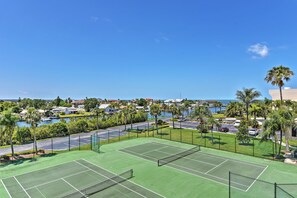 Un court de tennis avec un filet, entouré d’une clôture, et offrant une vue sur une étendue d’eau, avec des bâtiments et des palmiers en arrière-plan.