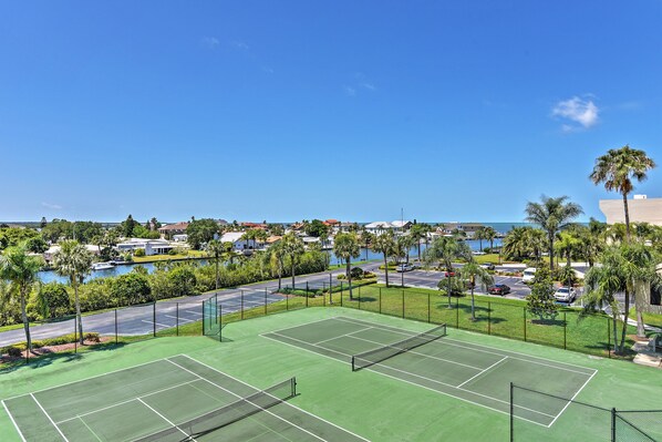 A tennis court with a net, surrounded by a fence, and overlooking a body of water with buildings and palm trees in the background. - On-site Beach Access! Waterfront Hudson Condo (Hudson)