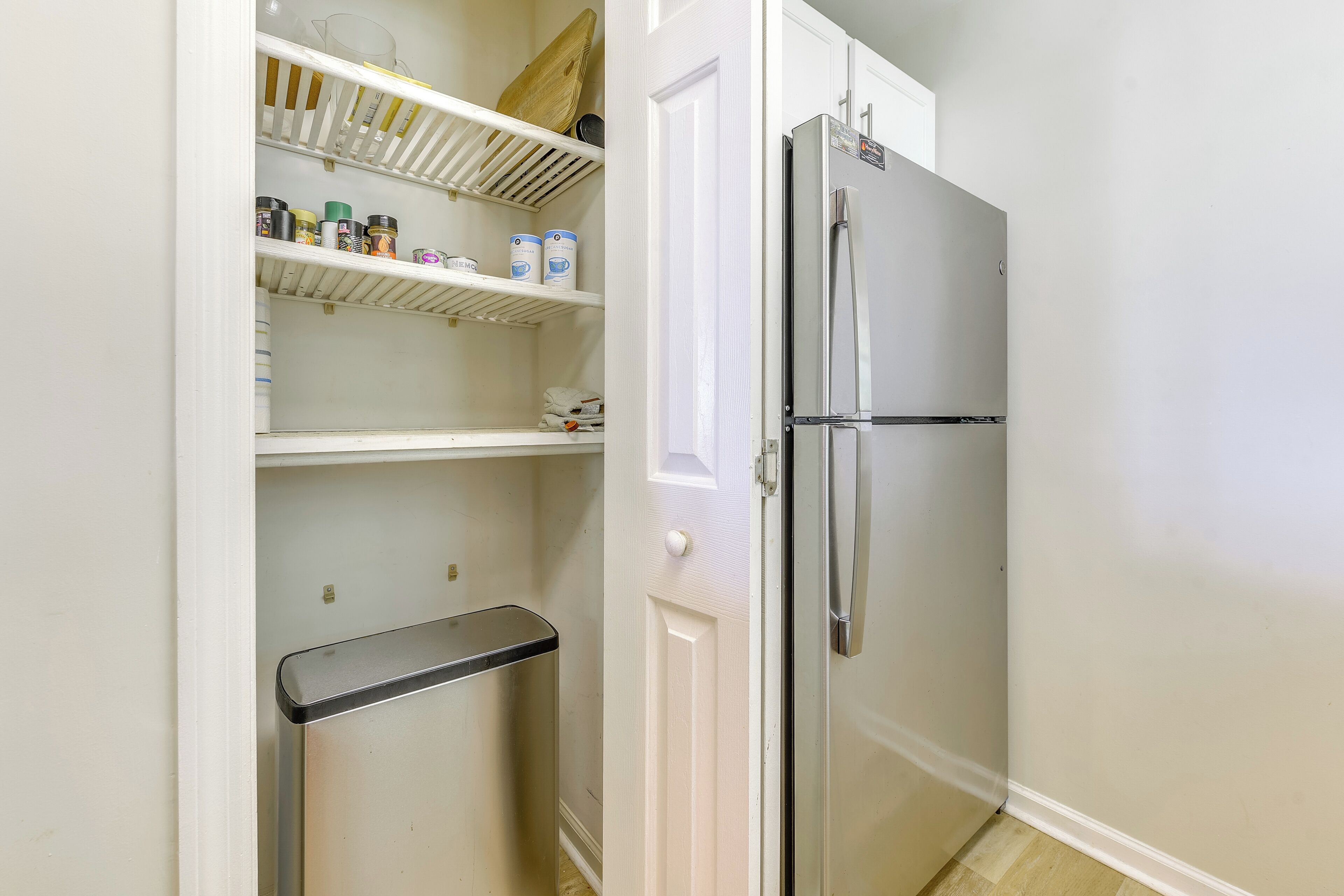 A kitchen with a stainless steel refrigerator, white shelves with various items, and a small countertop.