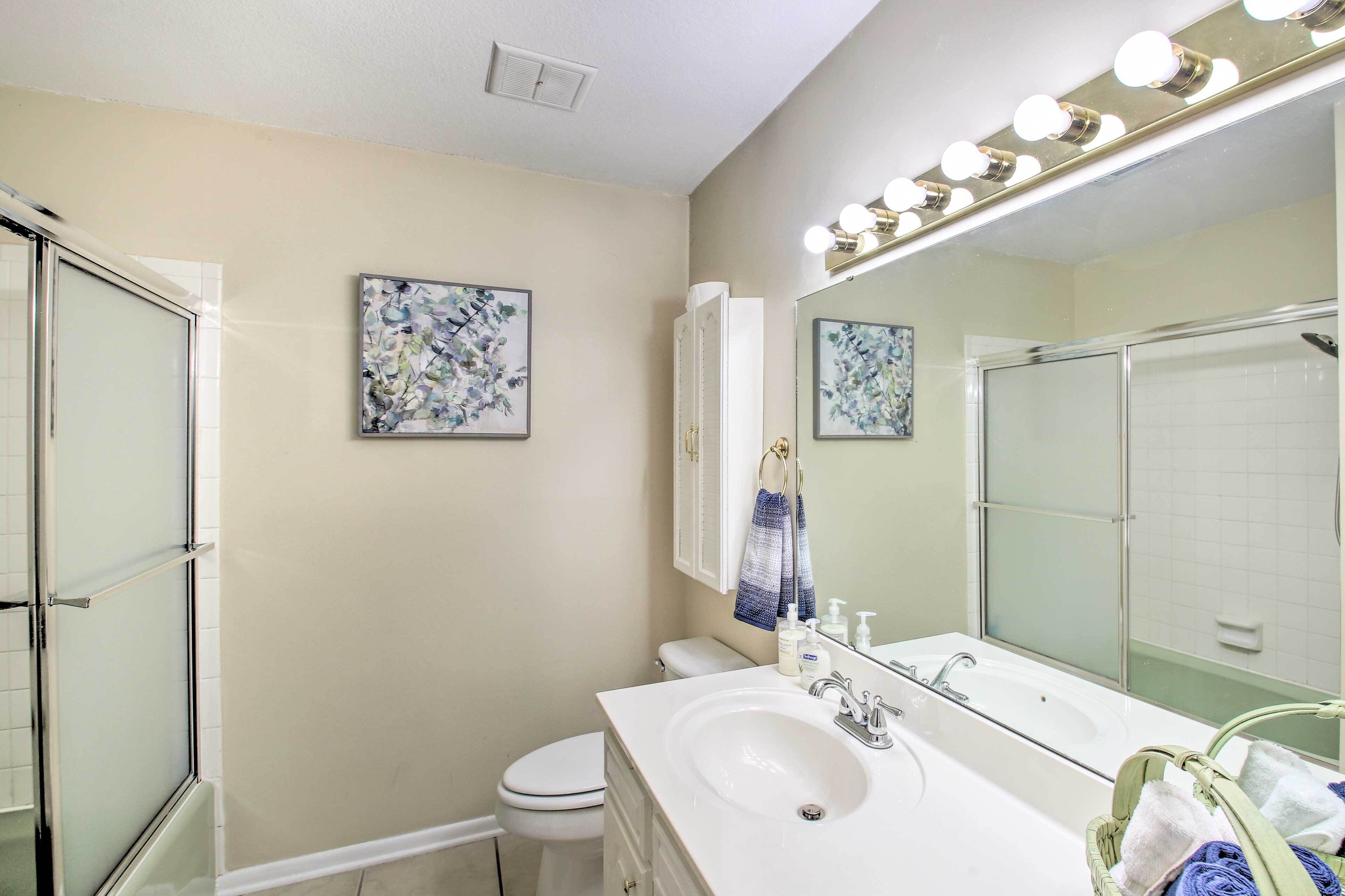 Un bagno con box doccia in vetro, un lavandino bianco, un ampio specchio con illuminazione e due quadri floreali incorniciati appesi al muro.
