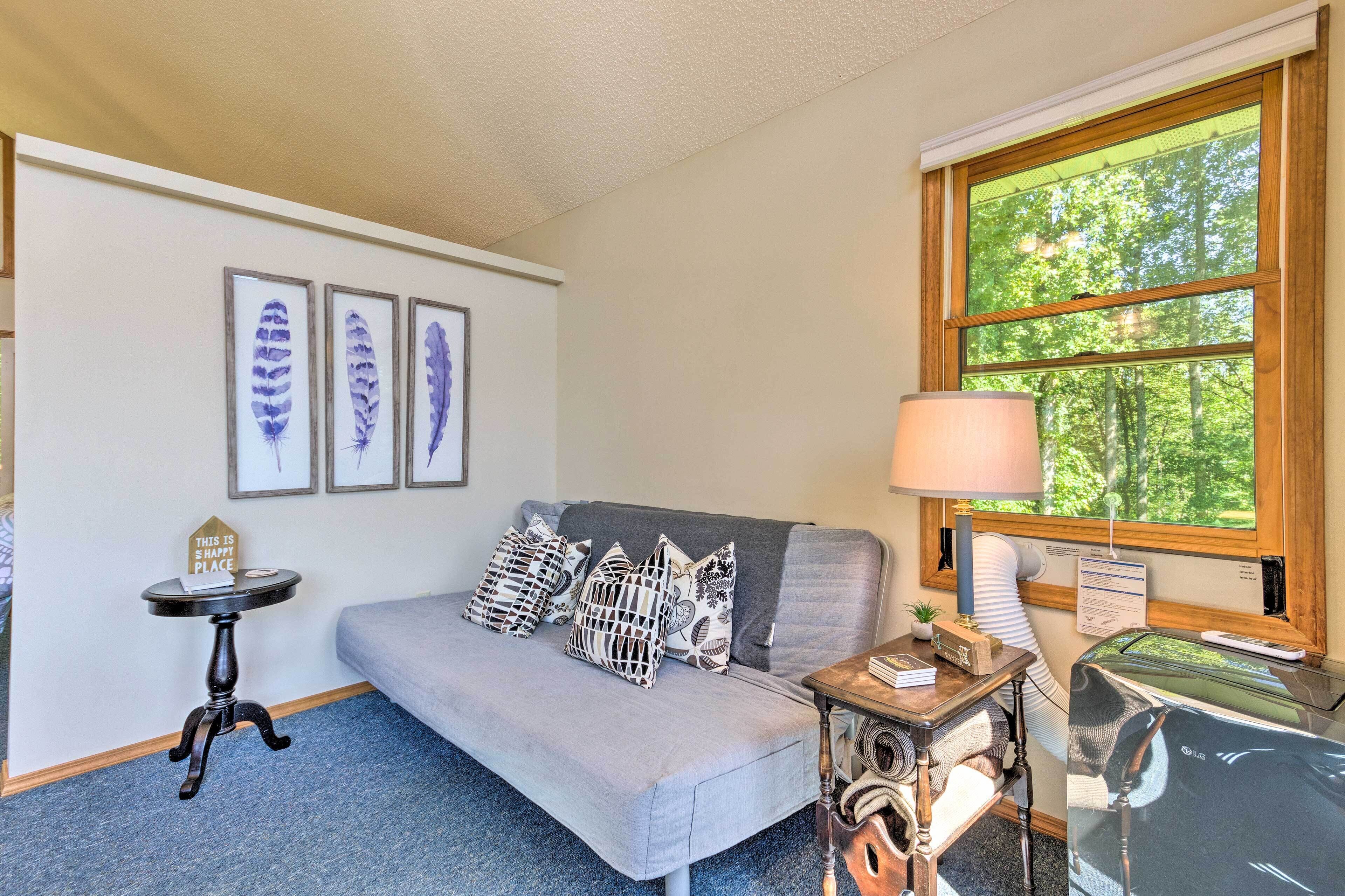 A cozy room with a grey sofa, a small table with a lamp, and framed feather art on the wall.