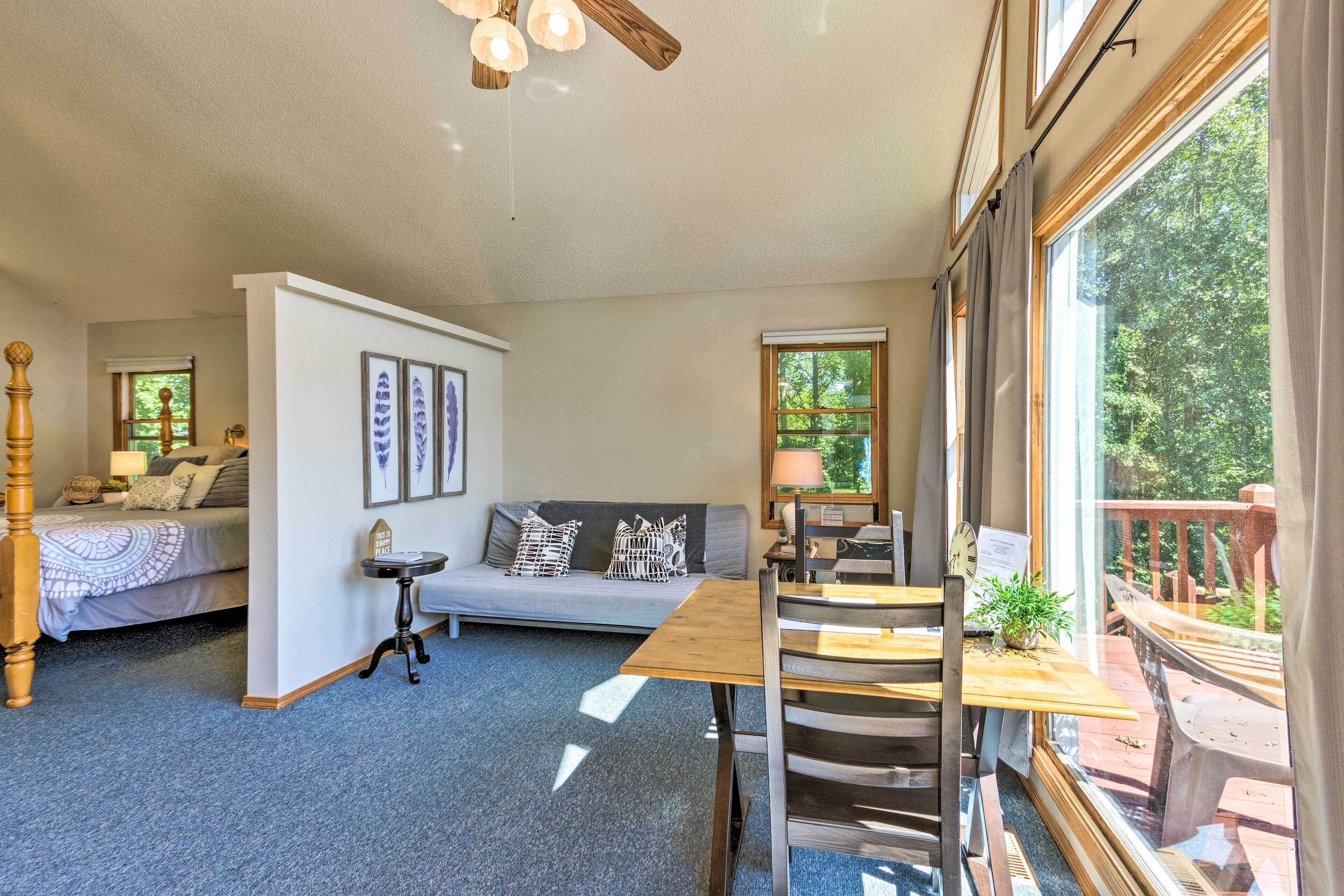 A cozy room with a bed, a sofa, a dining table, and a large window offering a view of the outdoors.