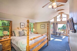 Un dormitorio amplio con una cama grande, cabecera de madera, ventilador de techo y vistas al exterior a través de amplias ventanas.