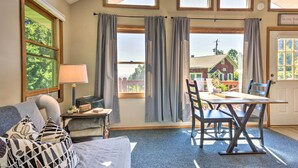 A room with a bed, a table with chairs, a lamp, and a view of a house through large windows. - Hendersonville 'cantrell Cottage' - Smoky Mtn View (Hendersonville)