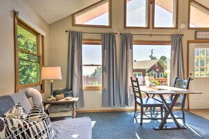 Una habitación con una cama, una mesa con sillas, una lámpara y vista a una casa a través de ventanas grandes.