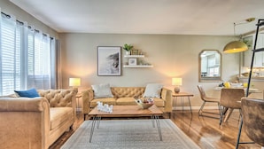 Ein modernes Wohnzimmer mit einem beigefarbenen Sofa, einem Couchtisch und einem Essbereich mit Stühlen. Ein Bücherregal mit gerahmten Bildern und einer Pflanze ist ebenfalls vorhanden.