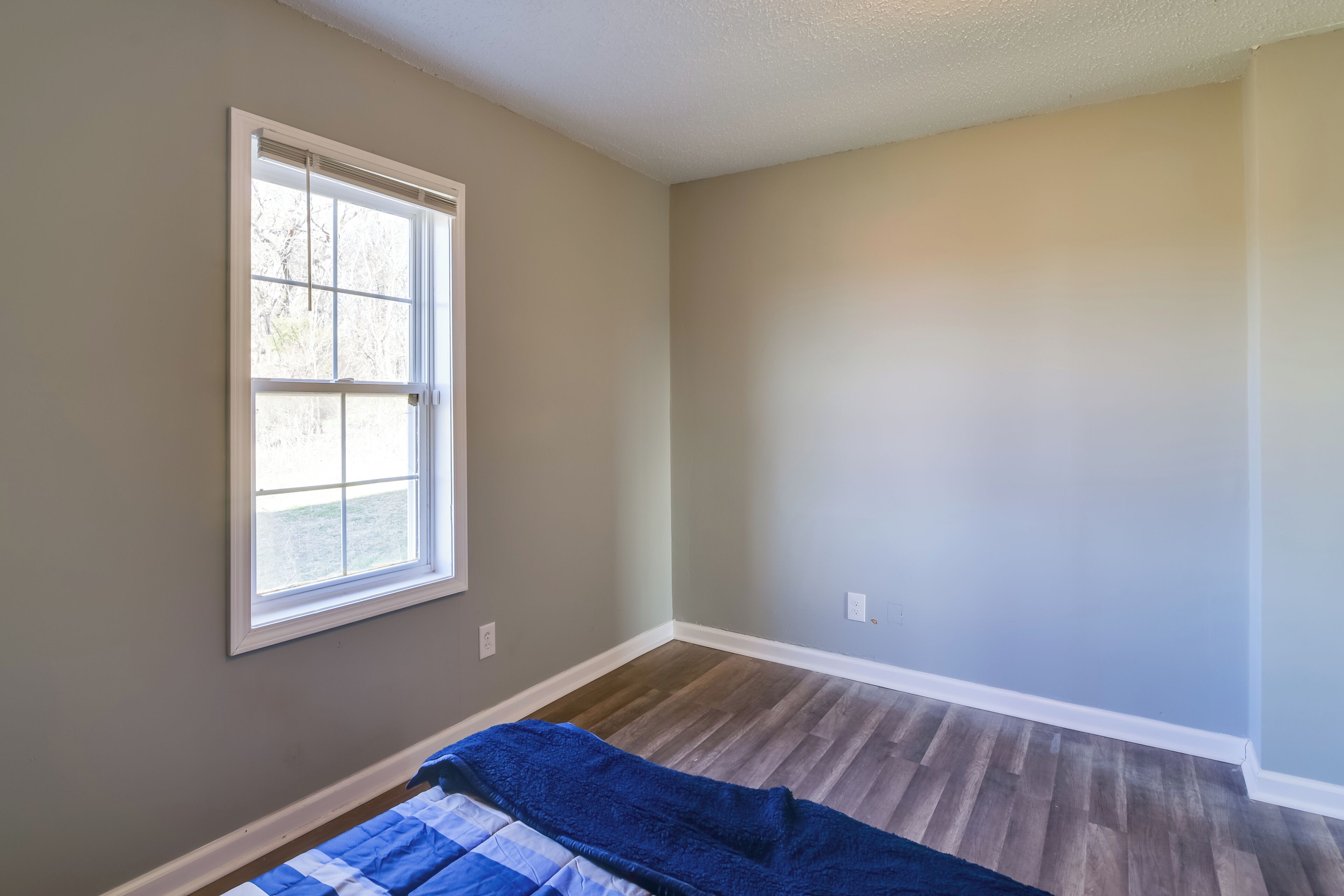 A room with a large window, wooden flooring, and a blue blanket on the bed.