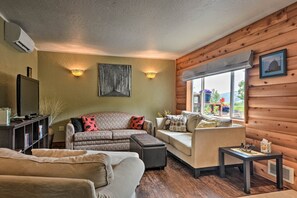 Ein gemütliches Wohnzimmer mit einer Couch, einem Hocker und einem kleinen Tisch. Ein Fernseher steht auf einem Lowboard, ein Fenster bietet einen Ausblick und eine Wandleuchte ist angebracht.