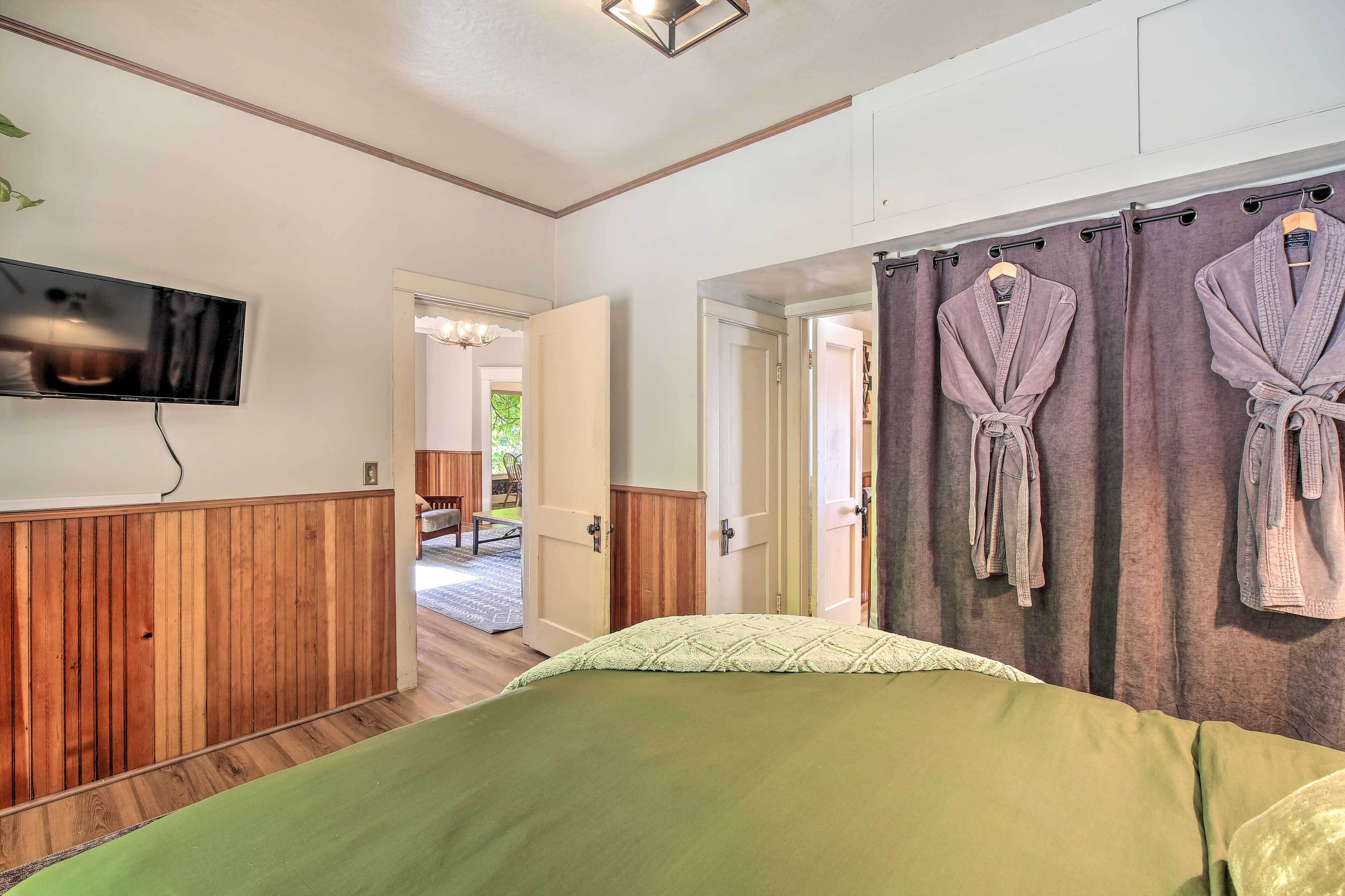 A bedroom with a green bed, wooden paneling, a TV mounted on the wall, and two robes hanging on the curtain rod.