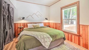 A bedroom with a wooden paneled wall, a large window with a view of trees, a bed with a green bedspread, and two wall-mounted lamps.