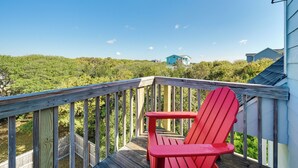 A wooden deck with a red Adirondack chair, overlooking a lush green landscape and a distant house.