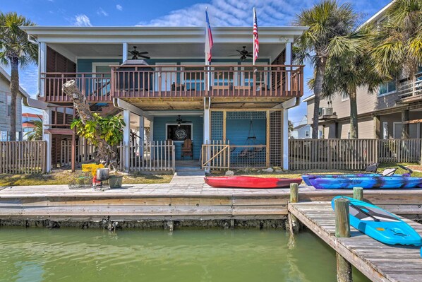 A waterfront property with a dock, kayaks, and a balcony with flags. - Jamaica Beach Waterfront Getaway: Kayaks & Dock! (Galveston)