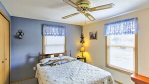 Une chambre à coucher comprenant un lit, un ventilateur de plafond, deux fenêtres avec des rideaux en dentelle, une table de chevet avec une lampe et un luminaire fixé au mur.