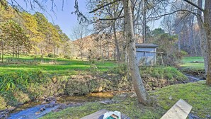 Blick auf einen Fluss mit einer Holzterrasse, einer kleinen Werkstatt und einem Picknicktisch mit Buch und Tasche.