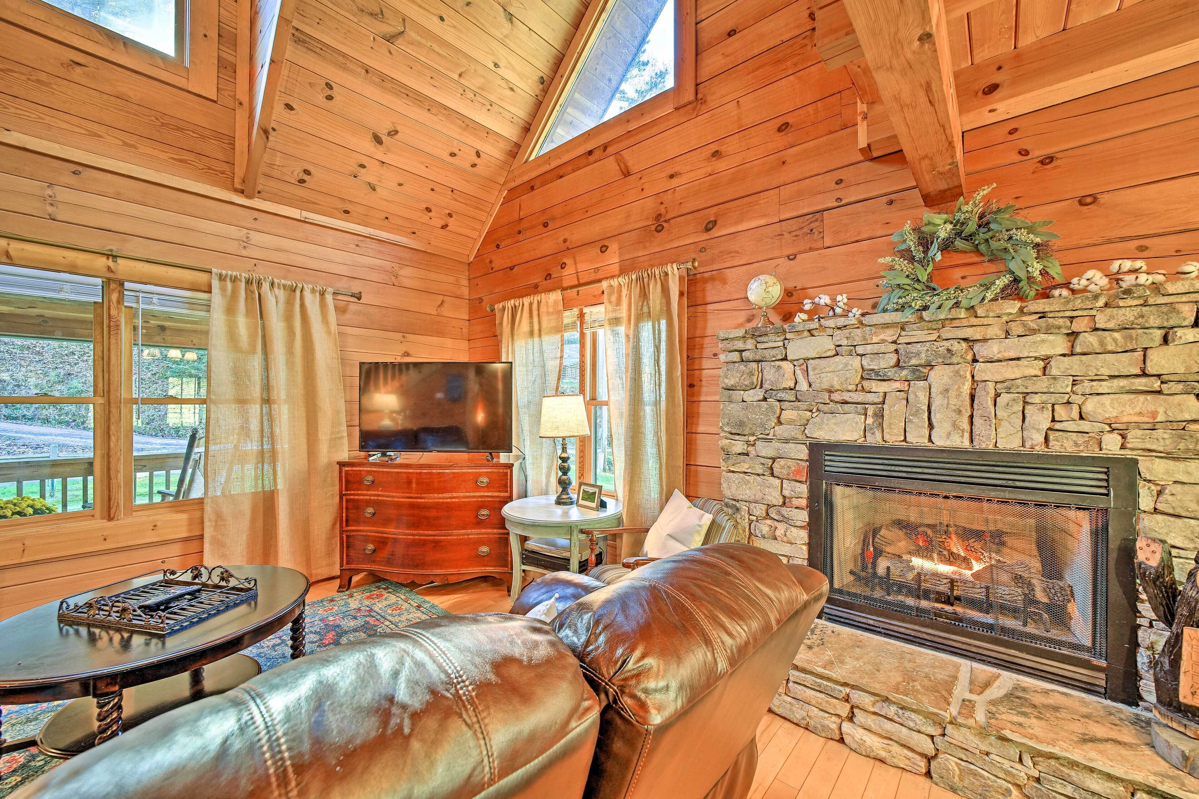 A cozy cabin interior with a stone fireplace, leather sofa, wooden walls, and a television.