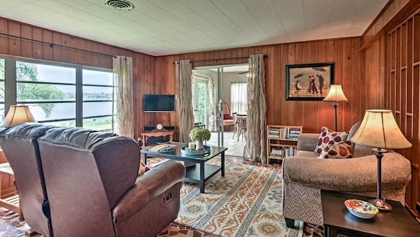 A cozy living room with a leather sofa, a glass coffee table, and a patterned rug. There is a television on a wooden stand, a lamp, and a painting on the wall. - Historic Lakefront Gem ~ 6 Mi to Bok Tower Gardens (Lake Wales)