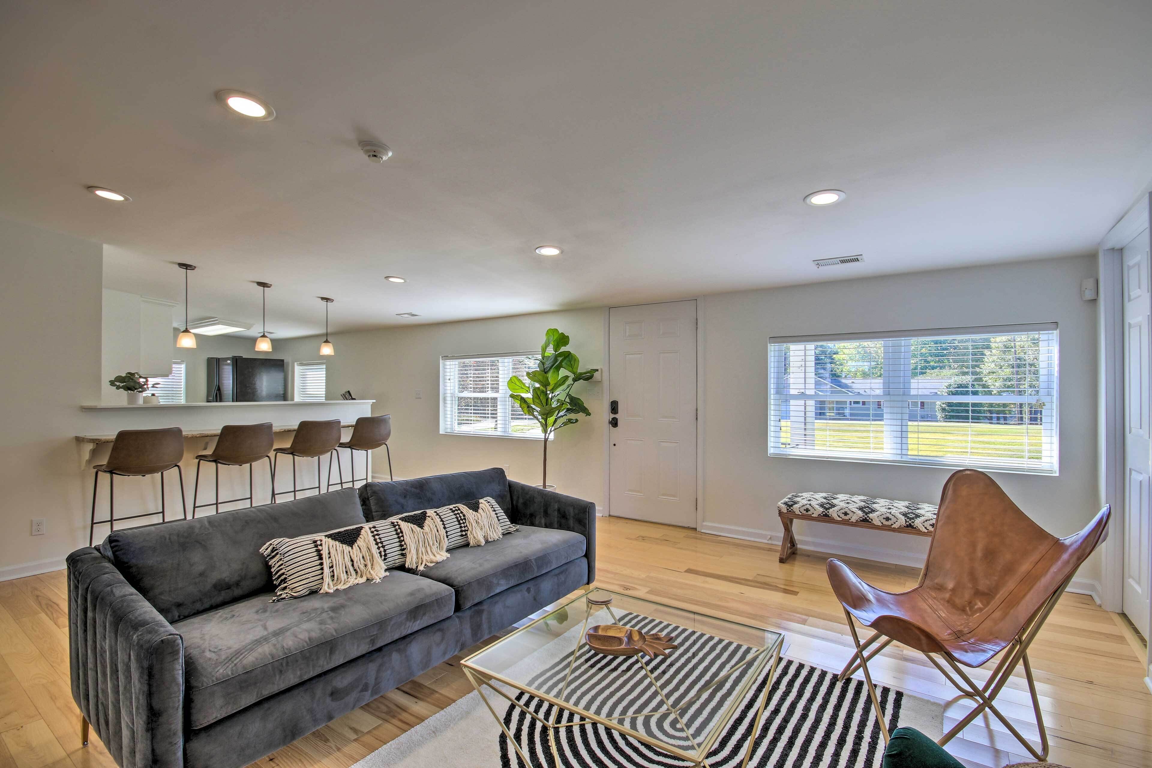 Una sala moderna con un sofá gris, una mesa de centro de cristal y un sillón de cuero. Hay una zona de comedor con taburetes altos y un banco.