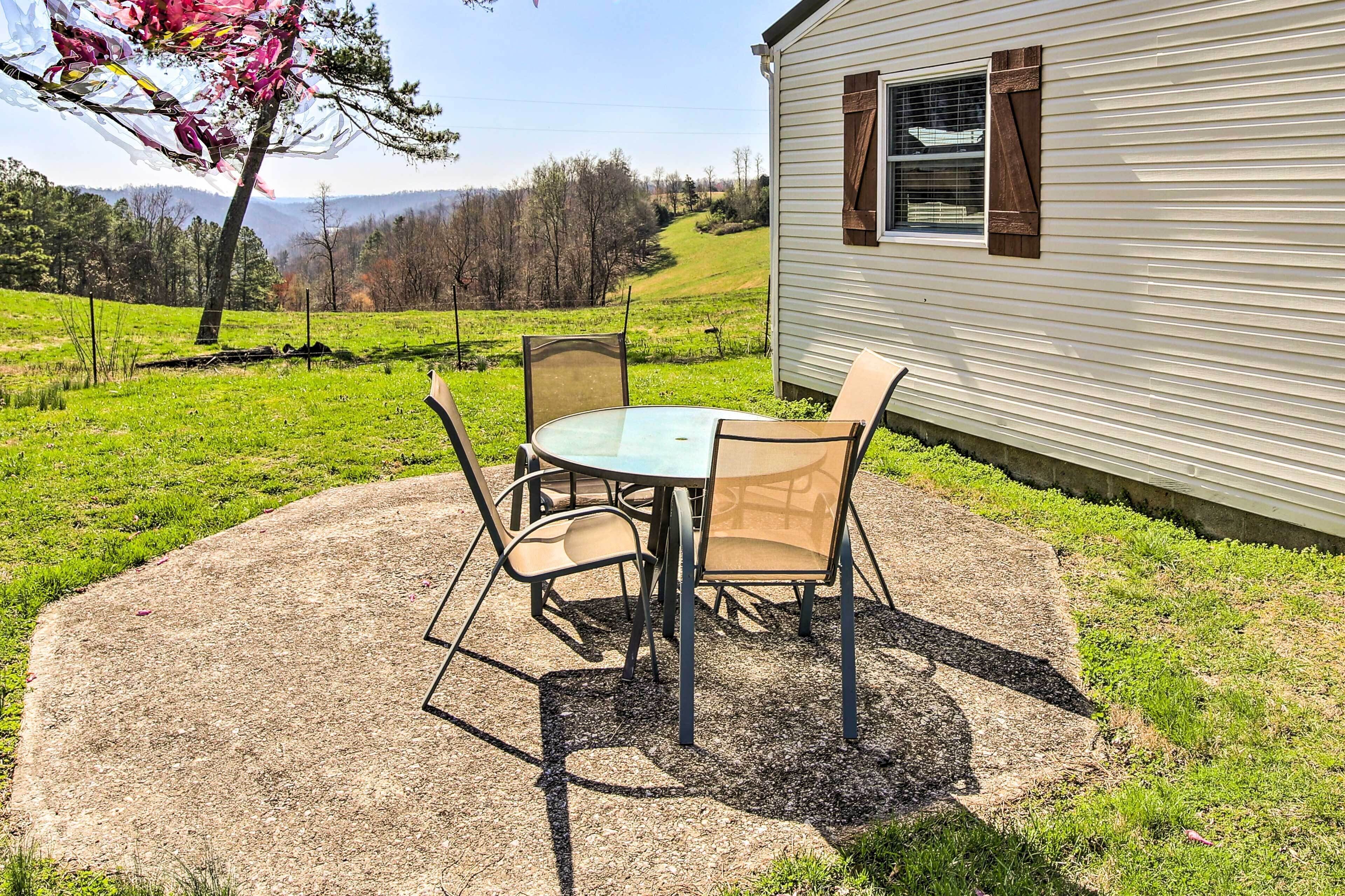Un patio con una mesa redonda y sillas, una casita con ventana, y un paisaje con césped, árboles y colinas al fondo.