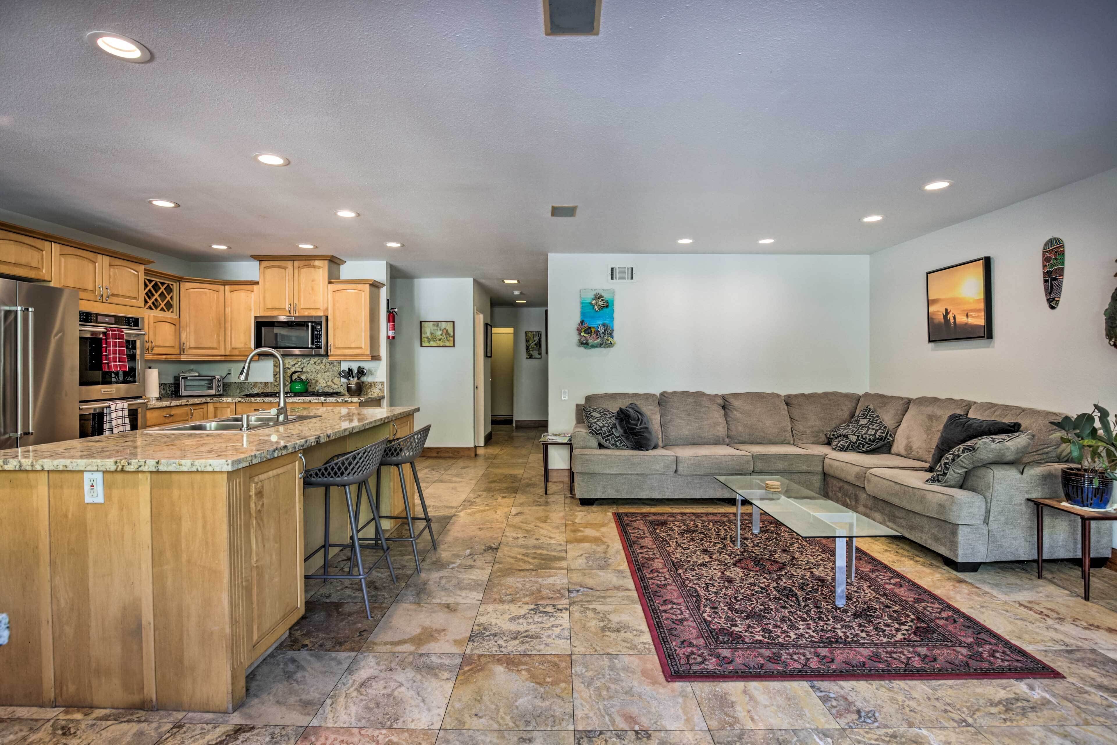 Cozinha moderna com ilha central, sala de jantar com sofá e mesa de centro, e sala de estar com um amplo sofá modular.