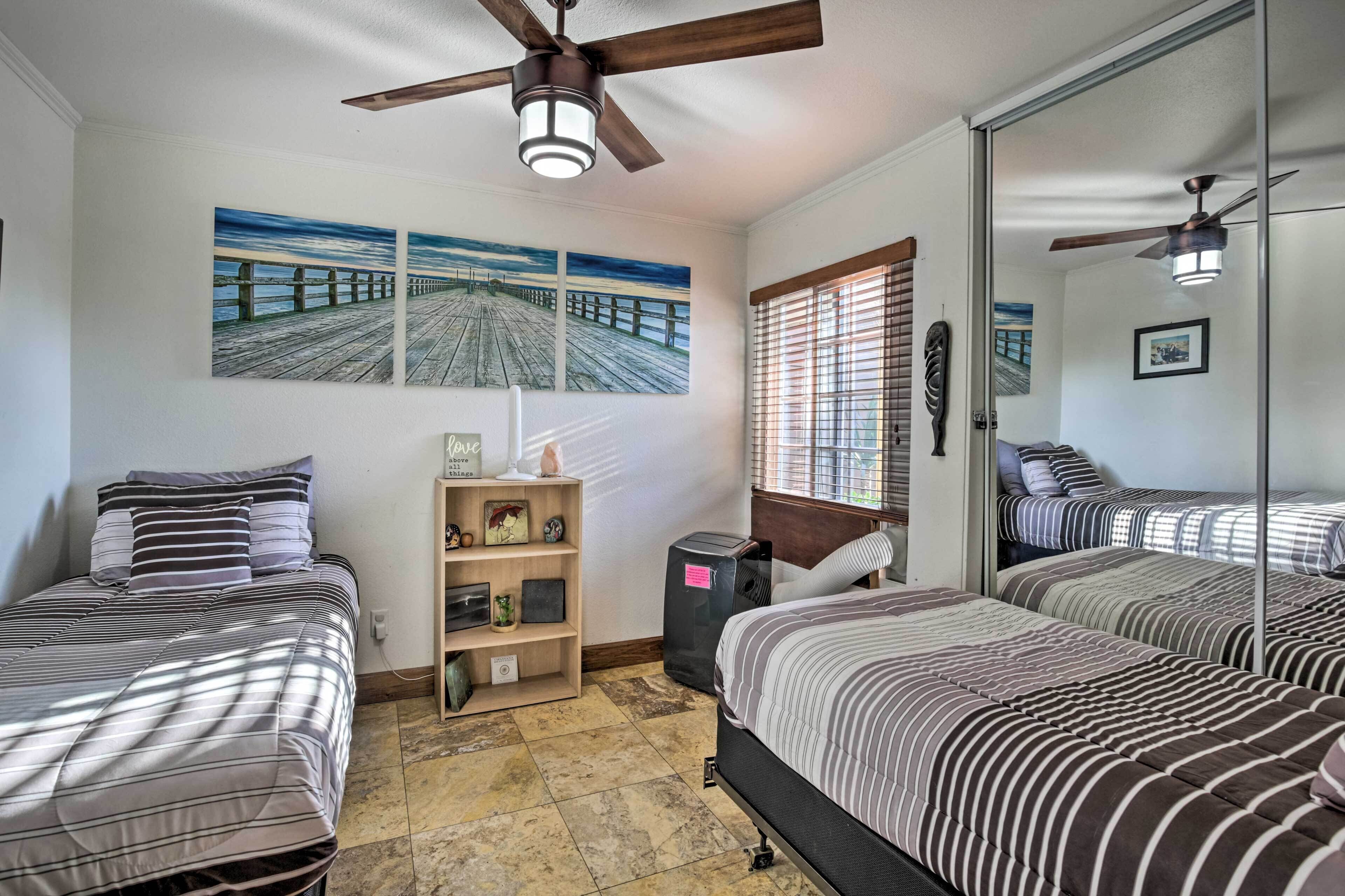 Quarto com duas camas, um ventilador de teto, uma prateleira com objetos decorativos e uma janela com persianas.
