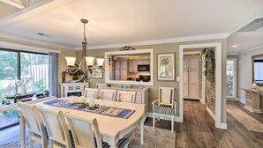 A dining area with a large table, chairs, and a chandelier. There is a kitchen area with cabinets and a microwave. The room has a wooden floor and a rug.