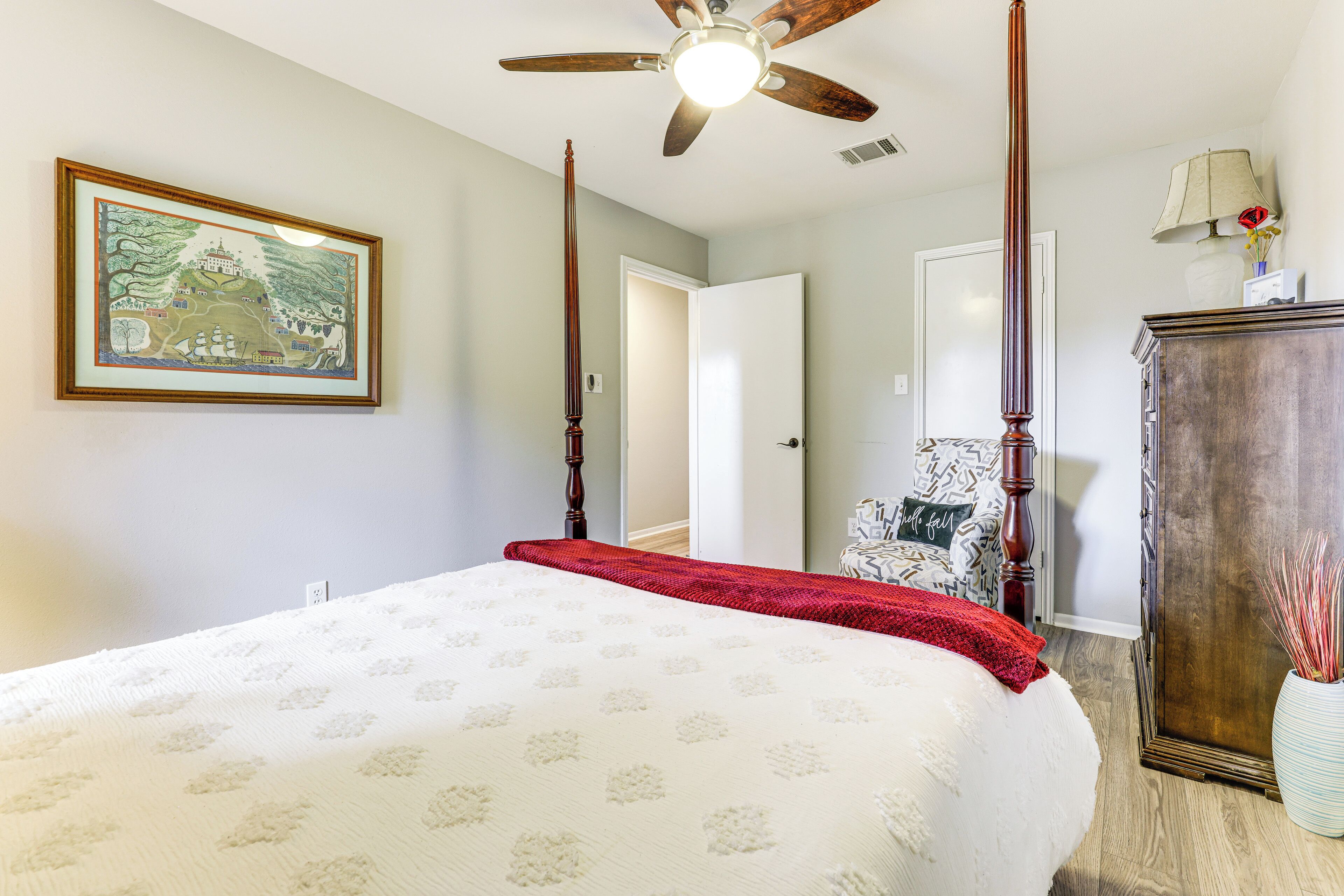 A bedroom with a four-poster bed, a ceiling fan, a framed painting, a wooden dresser, and a chair with a cushion.