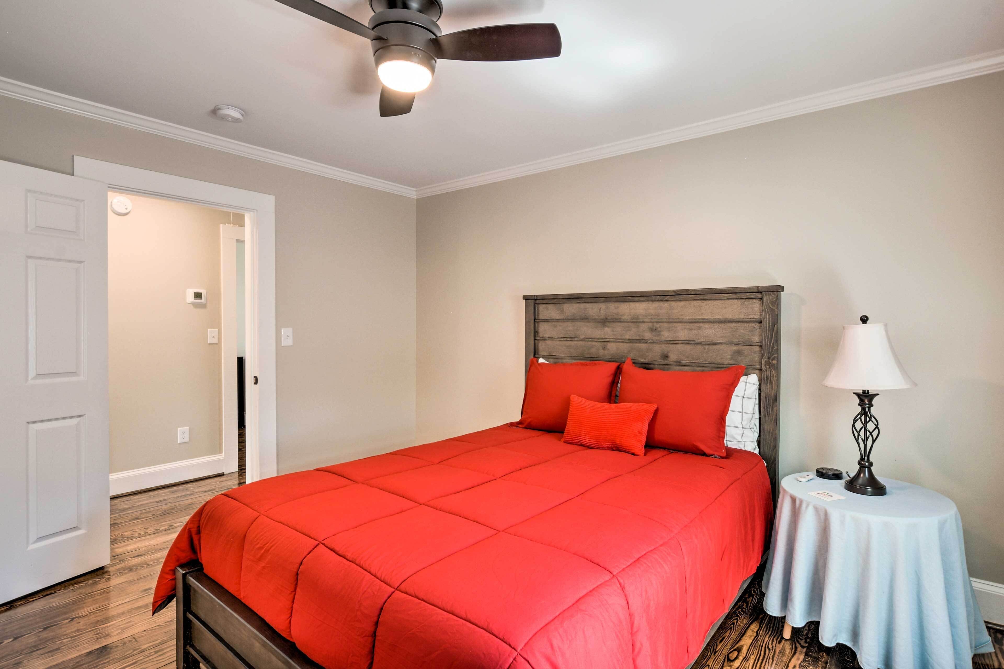 A bedroom with a large bed, a ceiling fan, a bedside table with a lamp, and a door leading to another room.