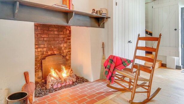 Ein Kamin mit Ziegelverkleidung, ein Holzschaukelstuhl mit kariertem Überwurf und ein Metallregal darüber.