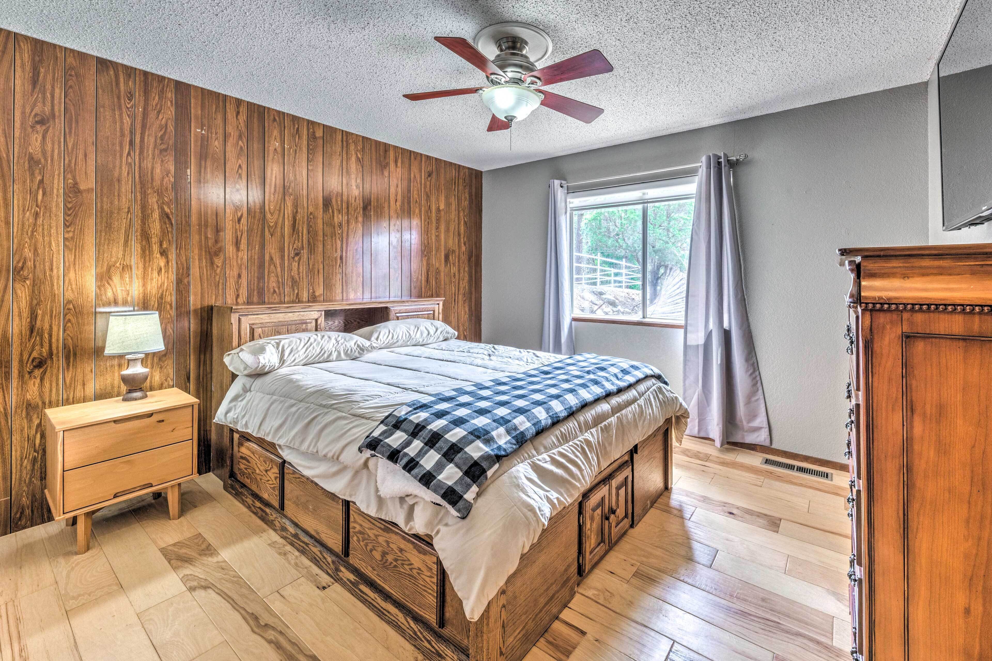 A bedroom with a wooden bed, a nightstand with a lamp, a ceiling fan, and a window with curtains.