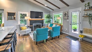 A modern living room with a fireplace, teal armchairs, a dining area with a long table, and a view of greenery through the windows.