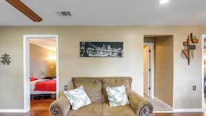 A living room with a brown sofa, two patterned cushions, a cityscape painting on the wall, and a wooden ceiling beam. - Kayak, Fish & Golf - Charming Victoria Home! (Bloomington)