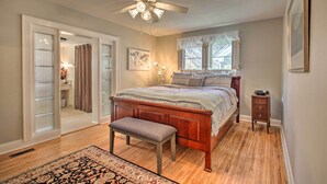 Quarto com cama de madeira, um banquinho, ventilador de teto e janela com cortinas.