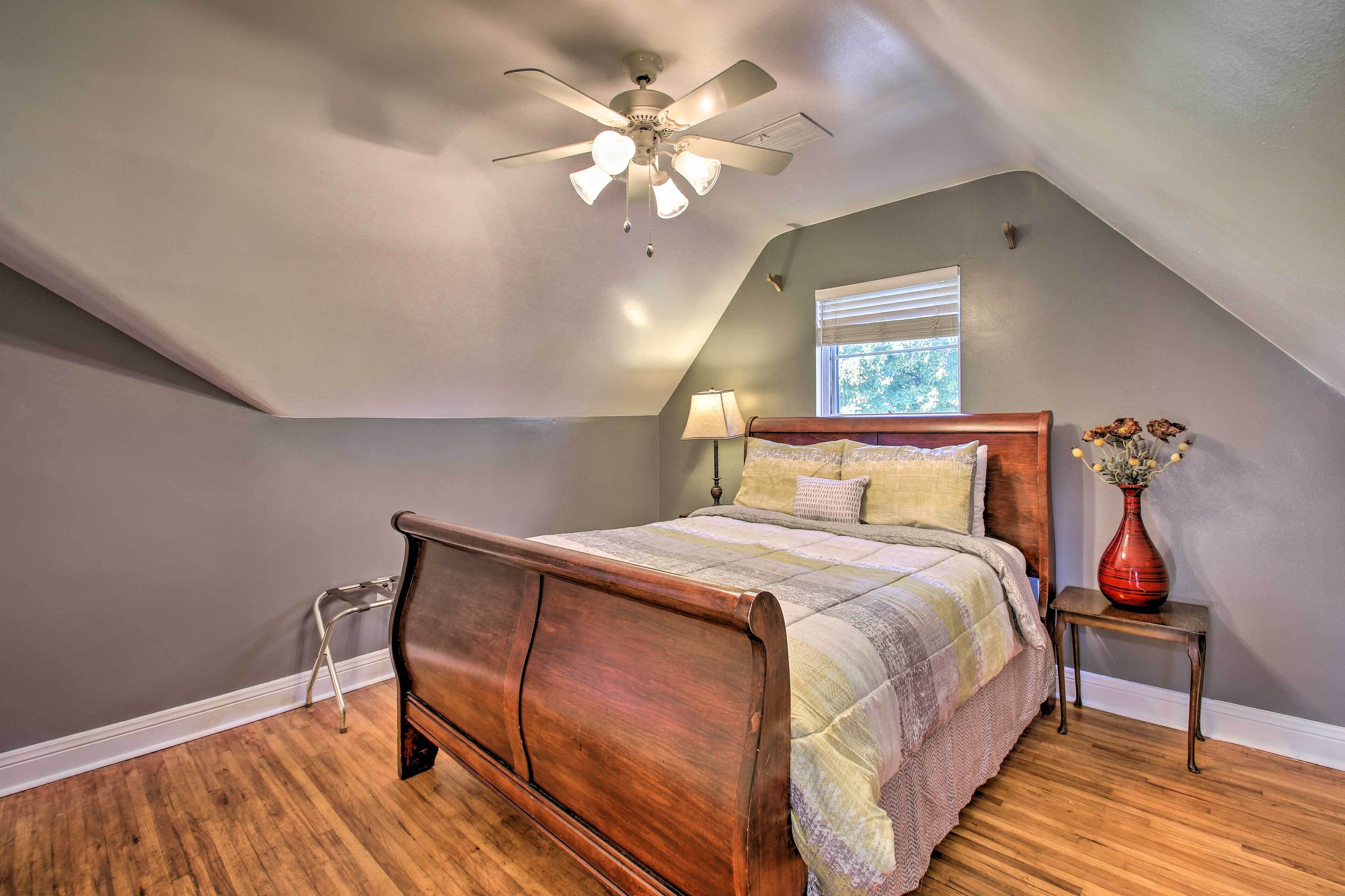 A bedroom with a wooden bed, a ceiling fan, a window with blinds, and a nightstand with a vase.