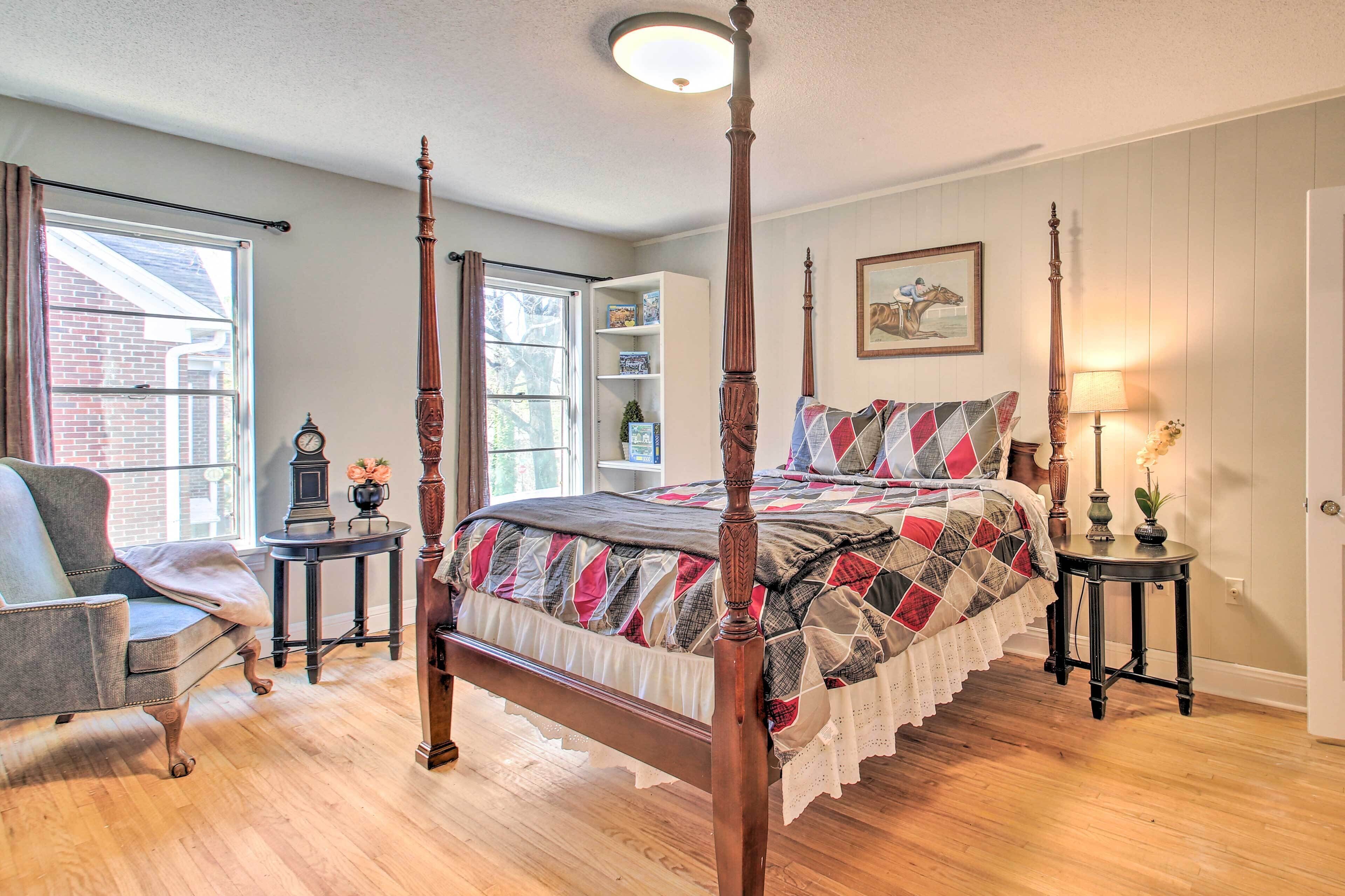 A four-poster bed with a quilt, a chair, a small table with a lamp, and a bookshelf.