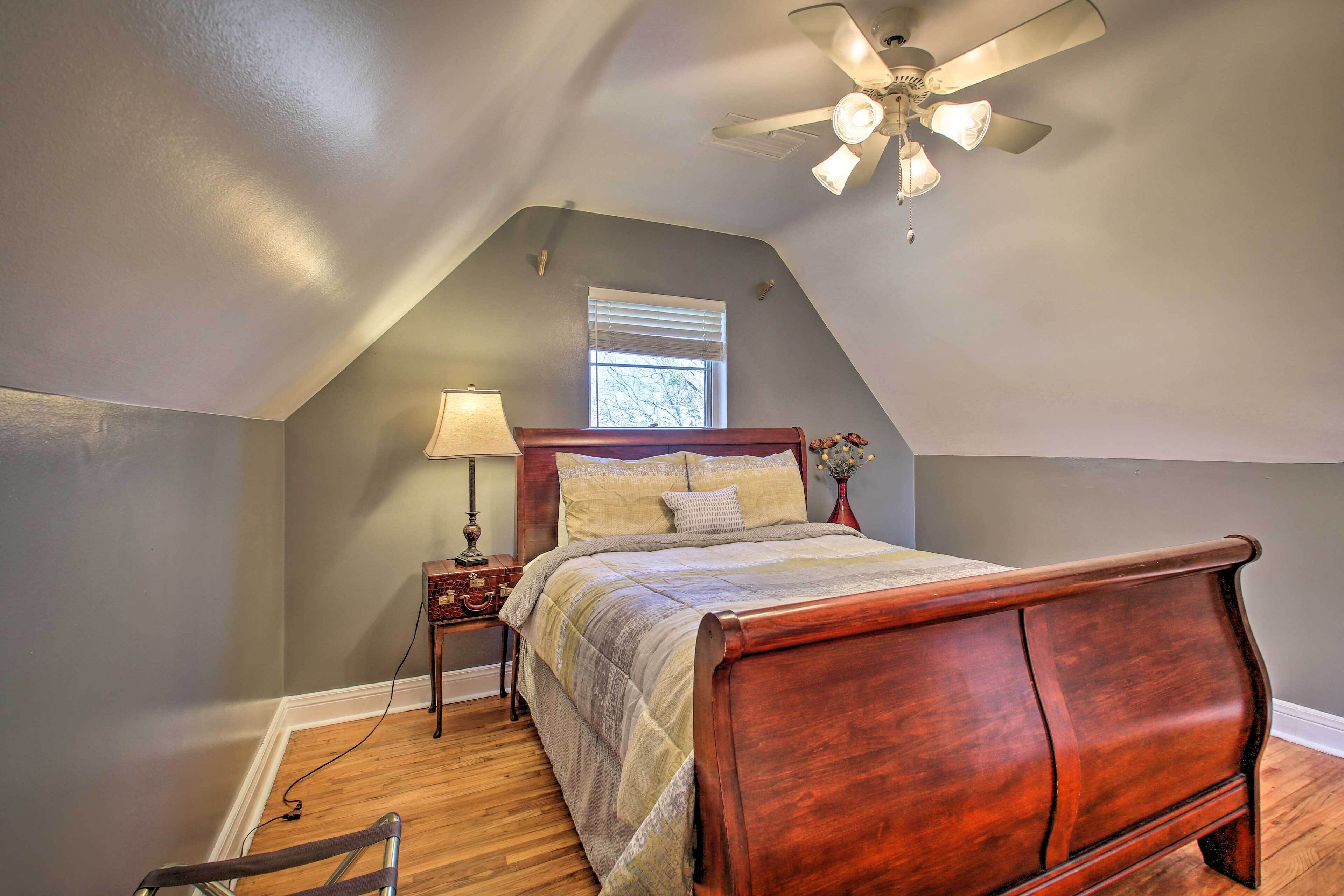 A bedroom with a sloped ceiling, a wooden bed, a nightstand, a lamp, and a window with blinds.