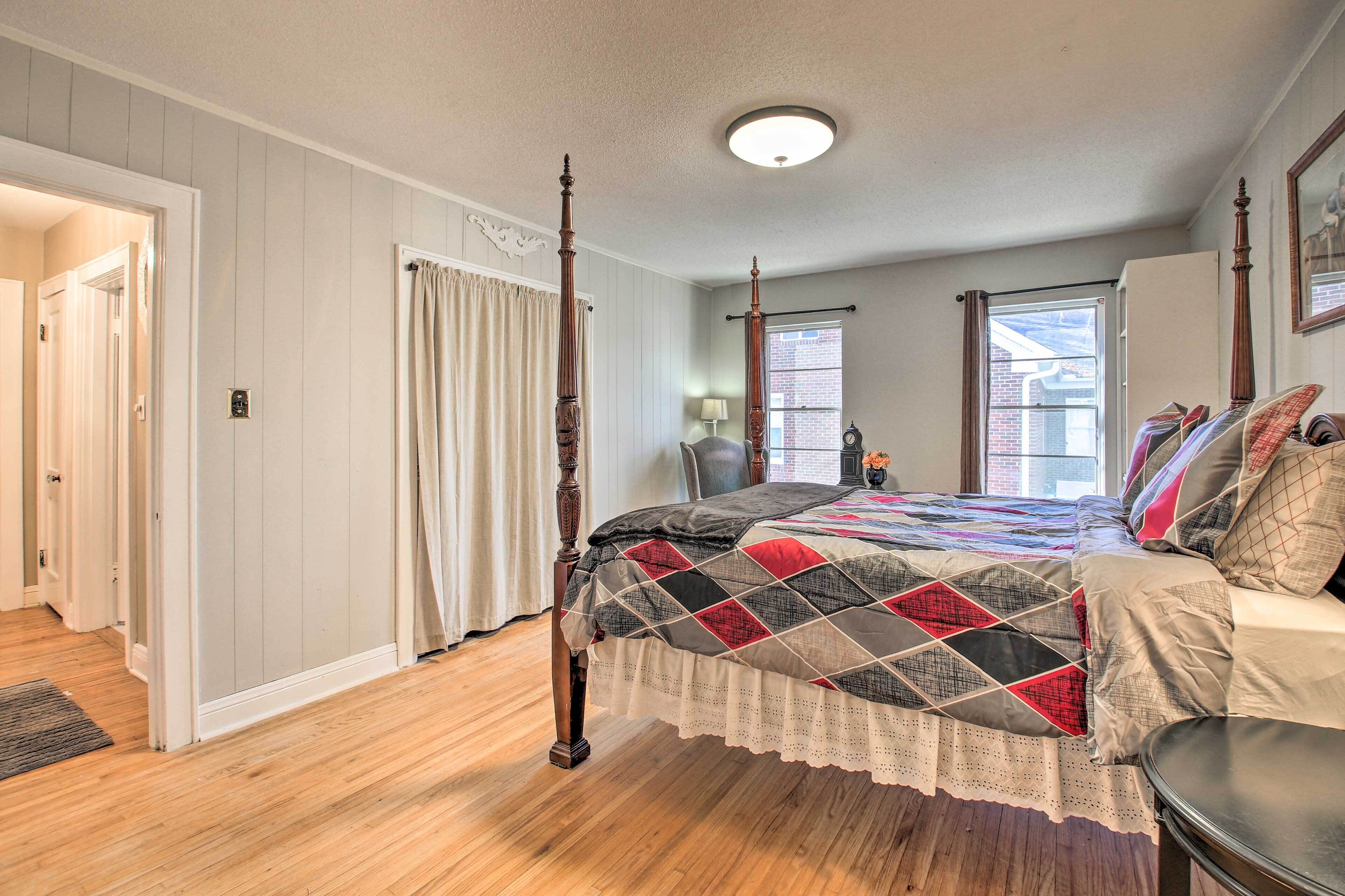 A four-poster bed with a quilt, a wooden floor, a window with blinds, and a door leading to another room.